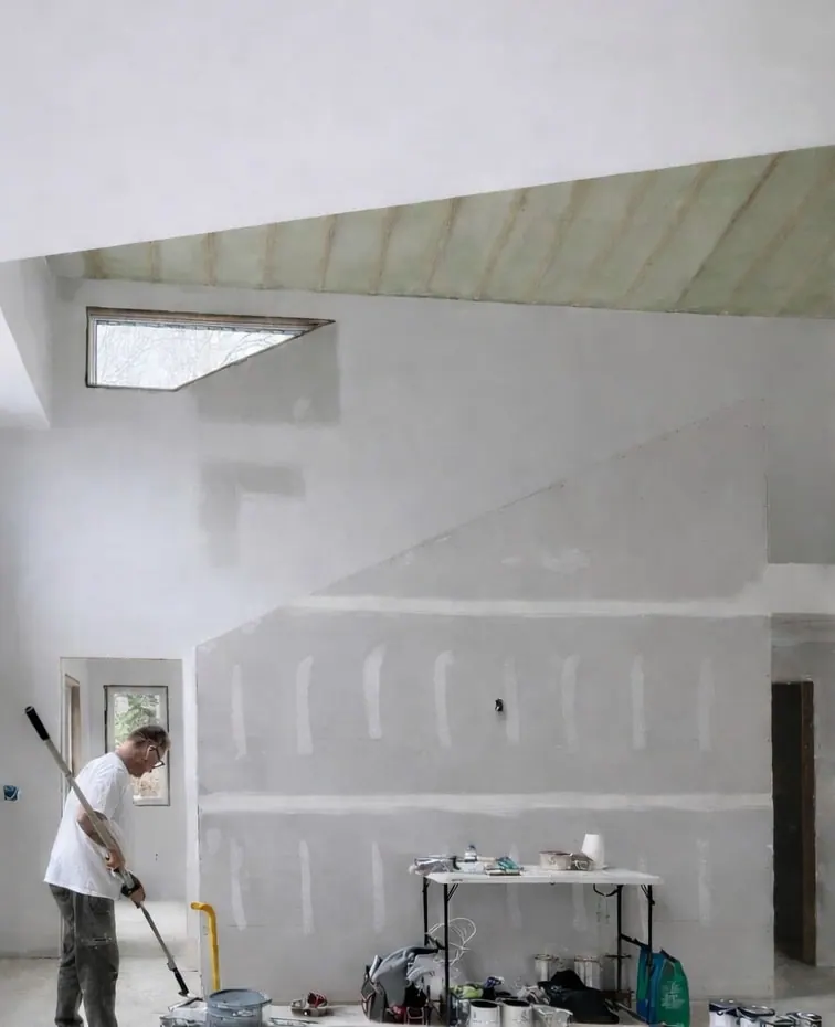 Unfinished drywall wall before painting, with a painter rolling primer